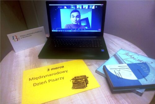 Na stoliku laptop, książki, kartka z napisem "3 marca Międzynarodowy Dzień Poezji" oraz logo Miejskiej Biblioteki Publicznej w Wasilkowie. Na ekranie laptopa mężczyzna.