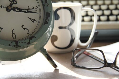 Stojący budzik, kubek i leżące okulary, w tle maszyna do pisania