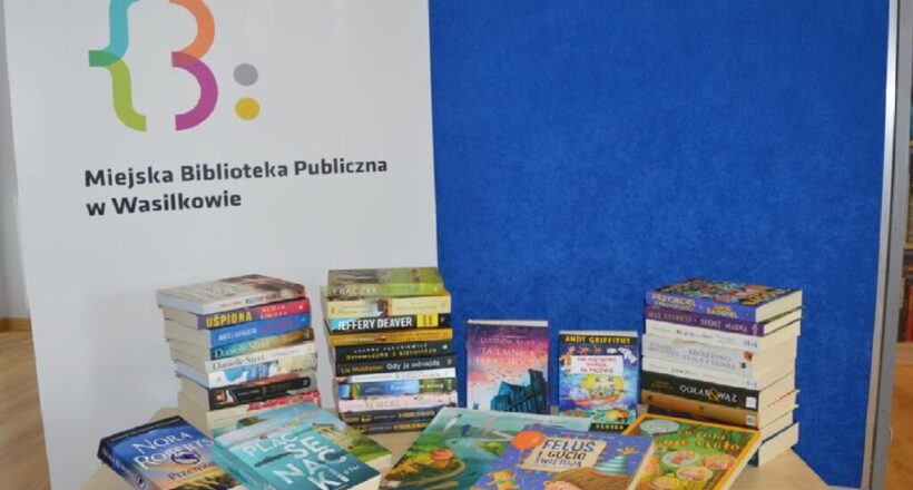 Książki wyeksponowane na tle loga Miejskiej Biblioteki Publicznej w Wasilkowie.