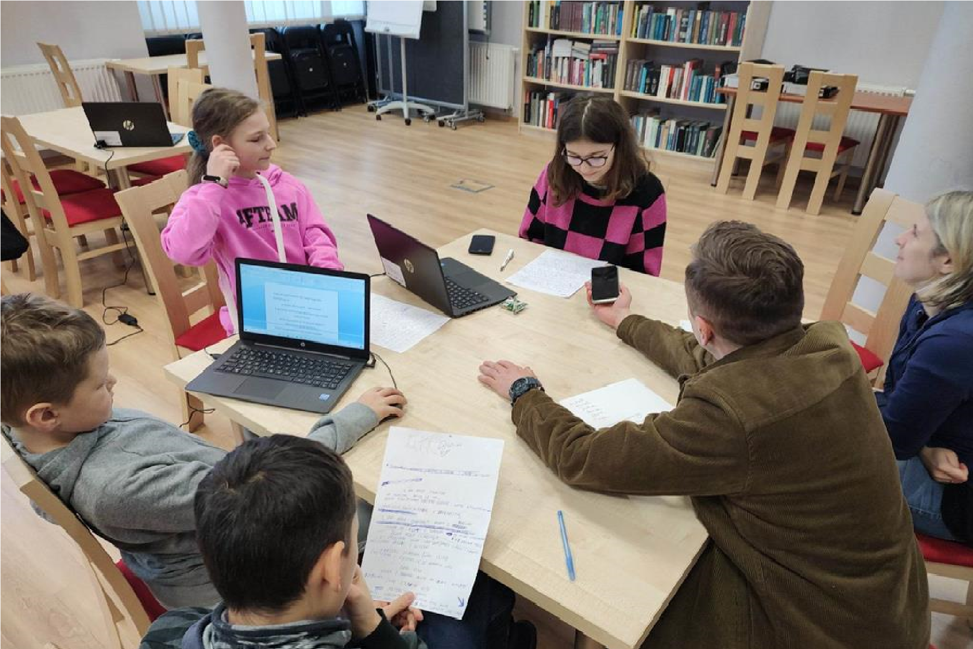 6 osób - dwóch chłopców, dwie dziewczyny, kobieta i mężczyzna siedzą przy stole w bibliotecznej czytelni. Na stole stoją dwa laptopy. Mężczyzna trzyma w ręku telefon i kieruje go w stronę jednej z dziewczyn.