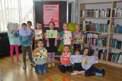 Grupa dzieci pozuje ze swoimi pracami plastycznymi na tle plakatu promującego DKK - Dyskusyjny Klub Książki.