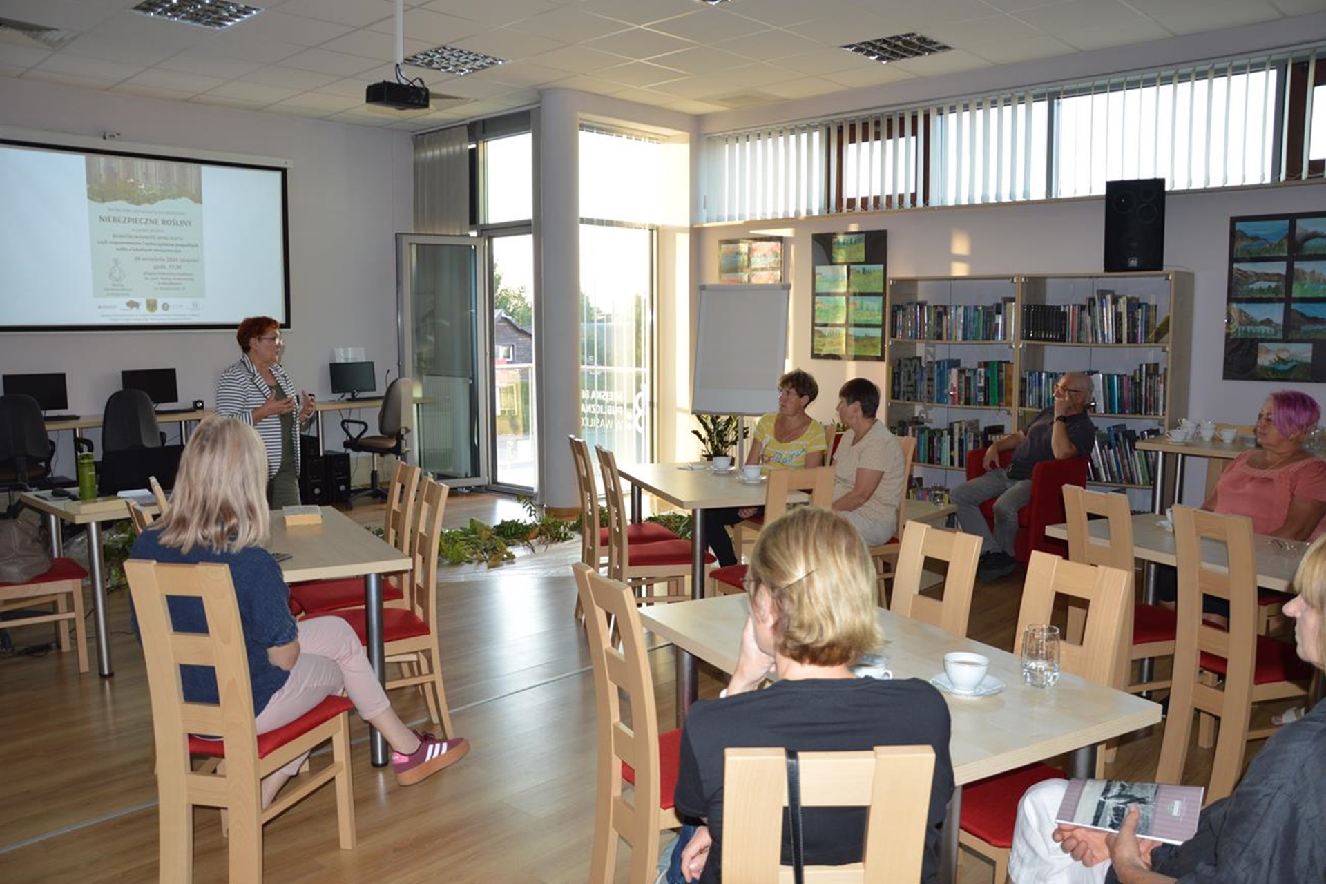 Grupa osób siedzi przy stolikach w bibliotecznej czytelni. Na środku sali stoi kobieta. W tle za nią widać otwarte drzwi balkonowe, stoły, krzesła, komputery i tablicę z wyświetlonym slajdem - plakatem promującym spotkanie "Niebezpieczne rośliny".