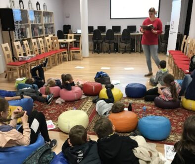 Kobieta czyta książkę siedzącej przed nią, na pufach, gromadce dzieci.